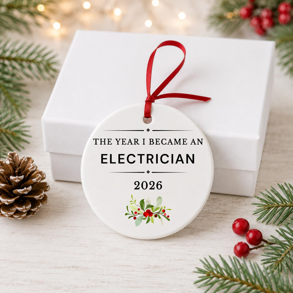 Electrician 2026 ceramic ornament reading The Year I Became an Electrician in front of a white gift box with pinecones and red berries
