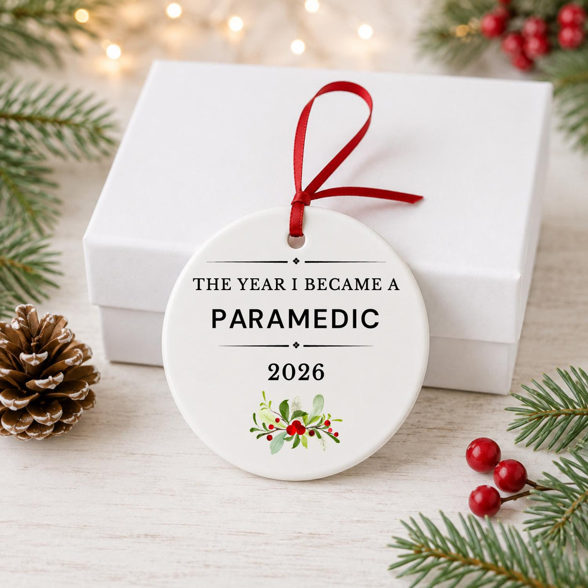 Paramedic 2026 ceramic ornament reading The Year I Became a Paramedic in front of a white gift box with pinecones and red berries