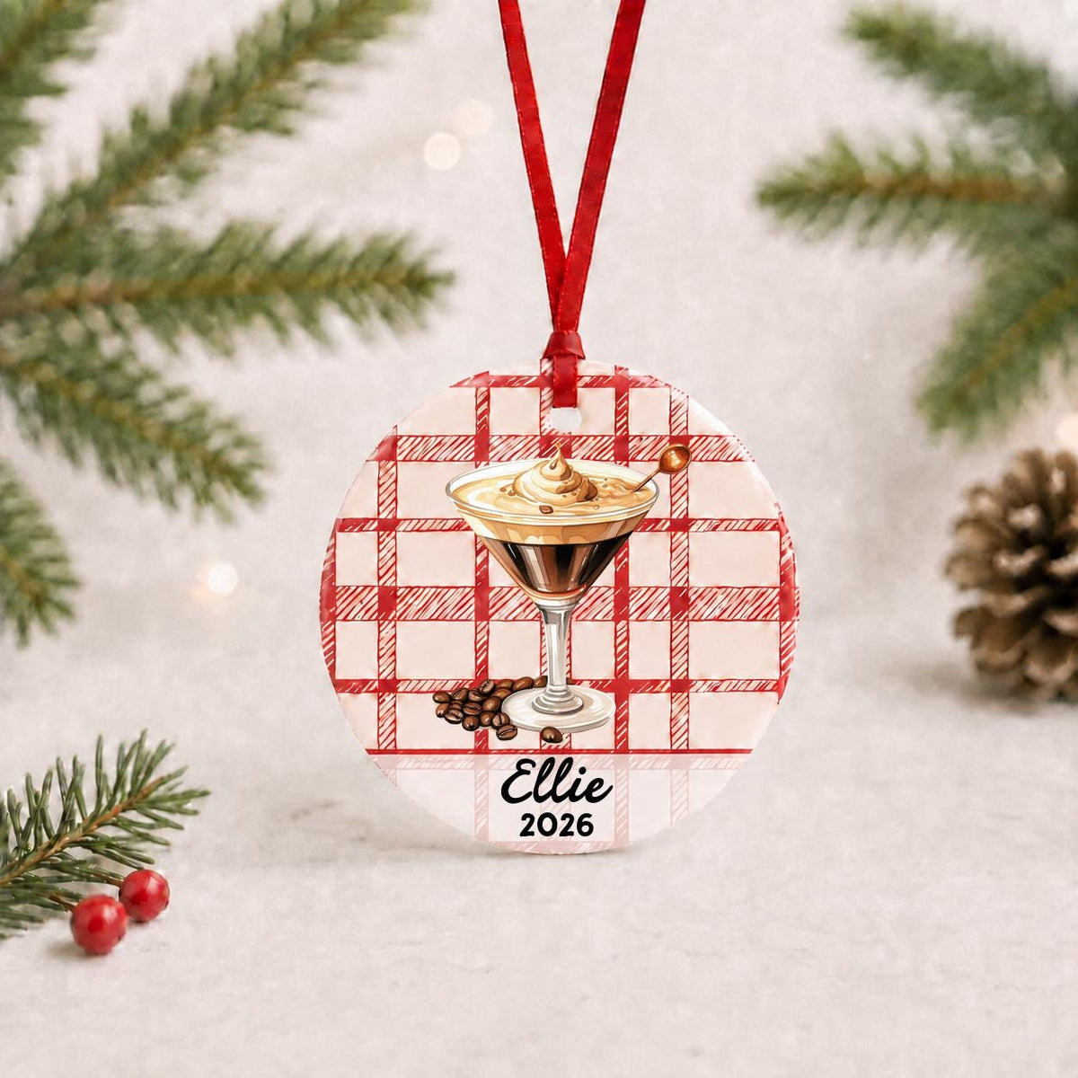 Personalized espresso martini ceramic ornament with name and year on red plaid with red ribbon on a white surface with pine branches and pinecones