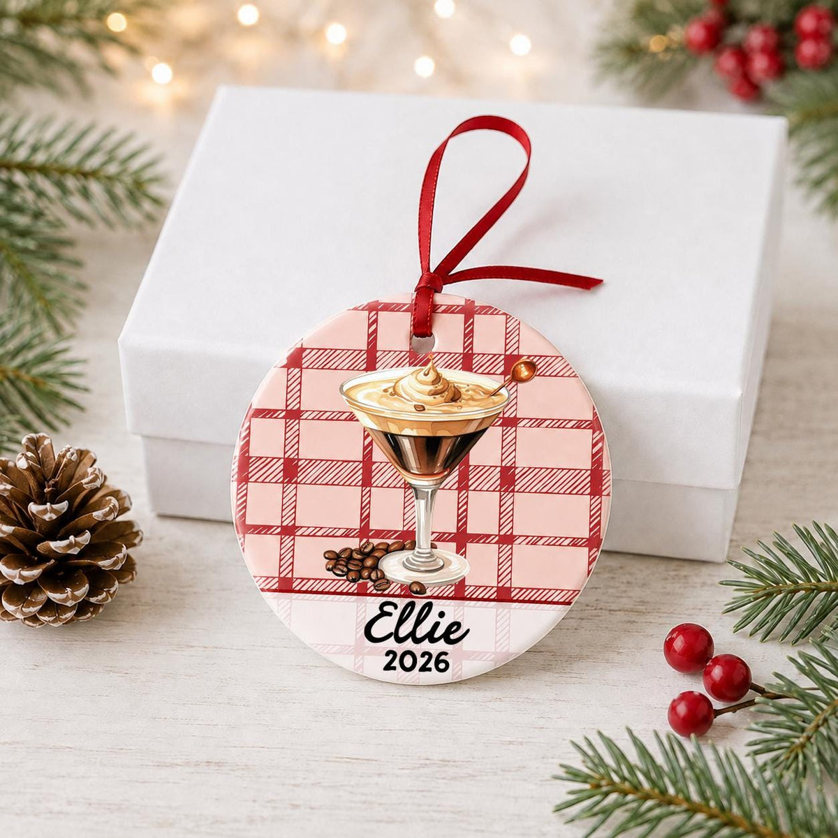 Personalized espresso martini ceramic ornament with name and year on red plaid in front of a white gift box with pinecones and red berries