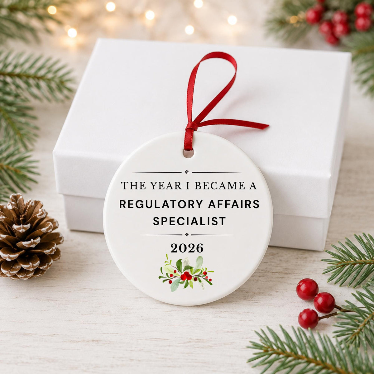 Regulatory Affairs Specialist 2026 ceramic ornament reading The Year I Became a Regulatory Affairs Specialist in front of a white gift box with pinecones and red berries