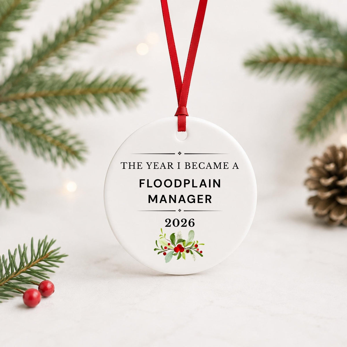 Floodplain Manager 2026 ceramic ornament reading The Year I Became a Floodplain Manager with red ribbon on a white surface with pine branches and pinecones