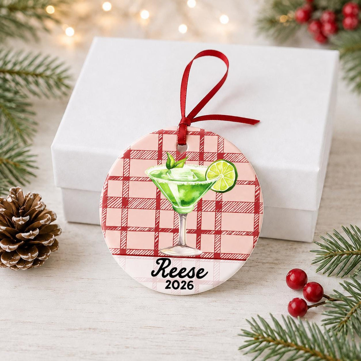 Personalized margarita ceramic ornament with name and year on red plaid in front of a white gift box with pinecones and red berries