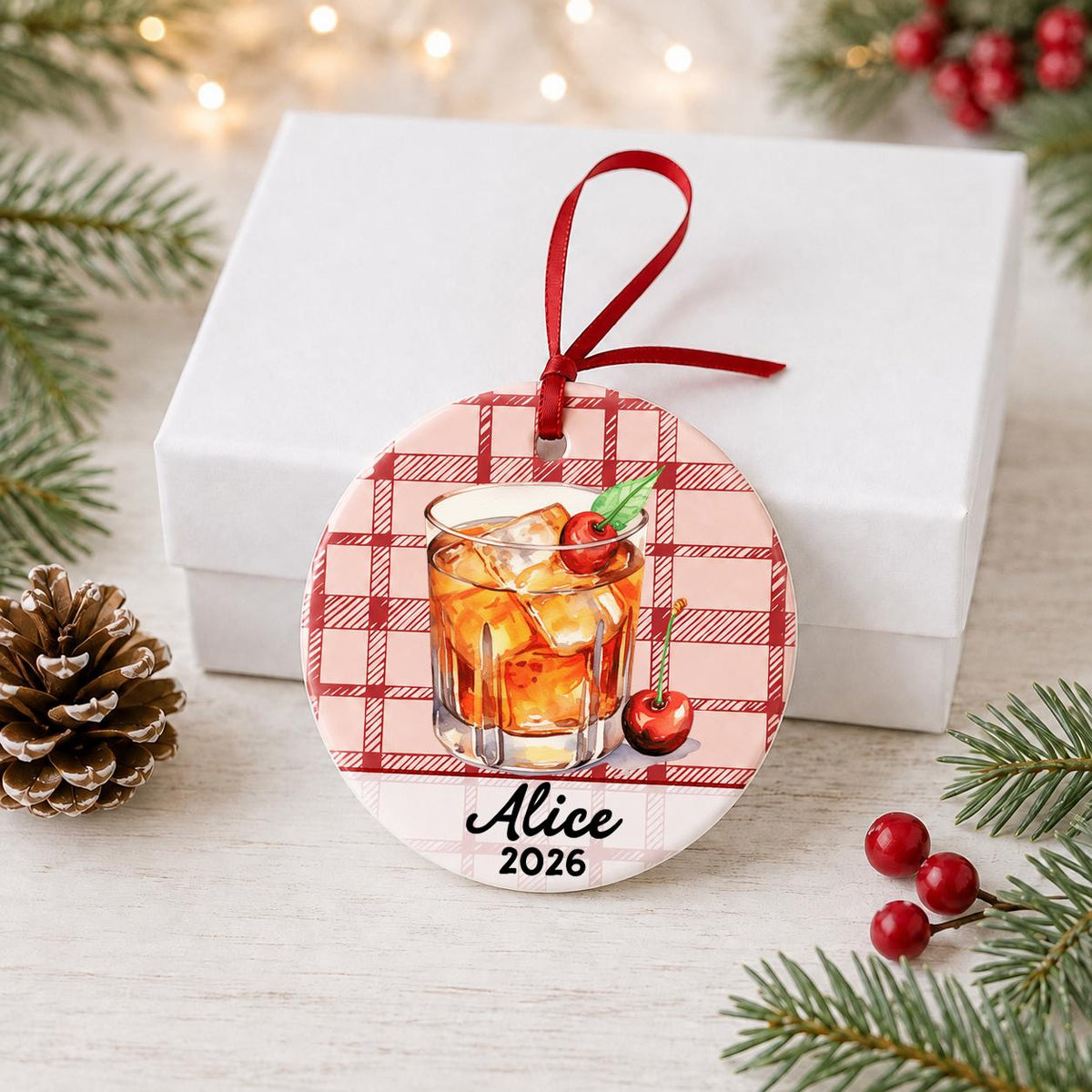 Personalized Old Fashioned cocktail ceramic ornament with name and year on red plaid in front of a white gift box with pinecones and red berries