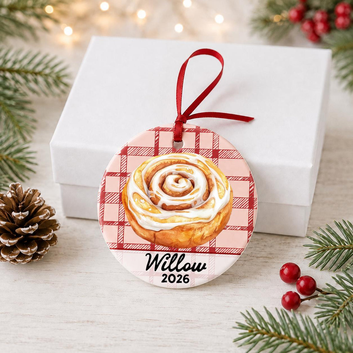 Personalized cinnamon roll ceramic ornament with name and year on red plaid in front of a white gift box with pinecones and red berries