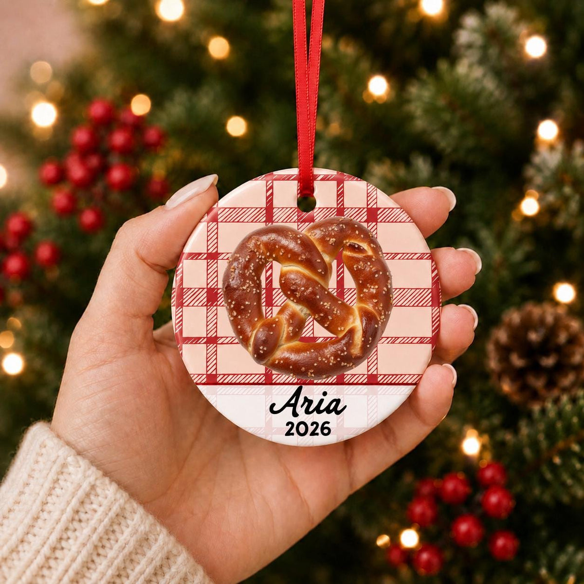 Hand holding the personalized soft pretzel ceramic ornament with name and year on red plaid in front of a Christmas tree