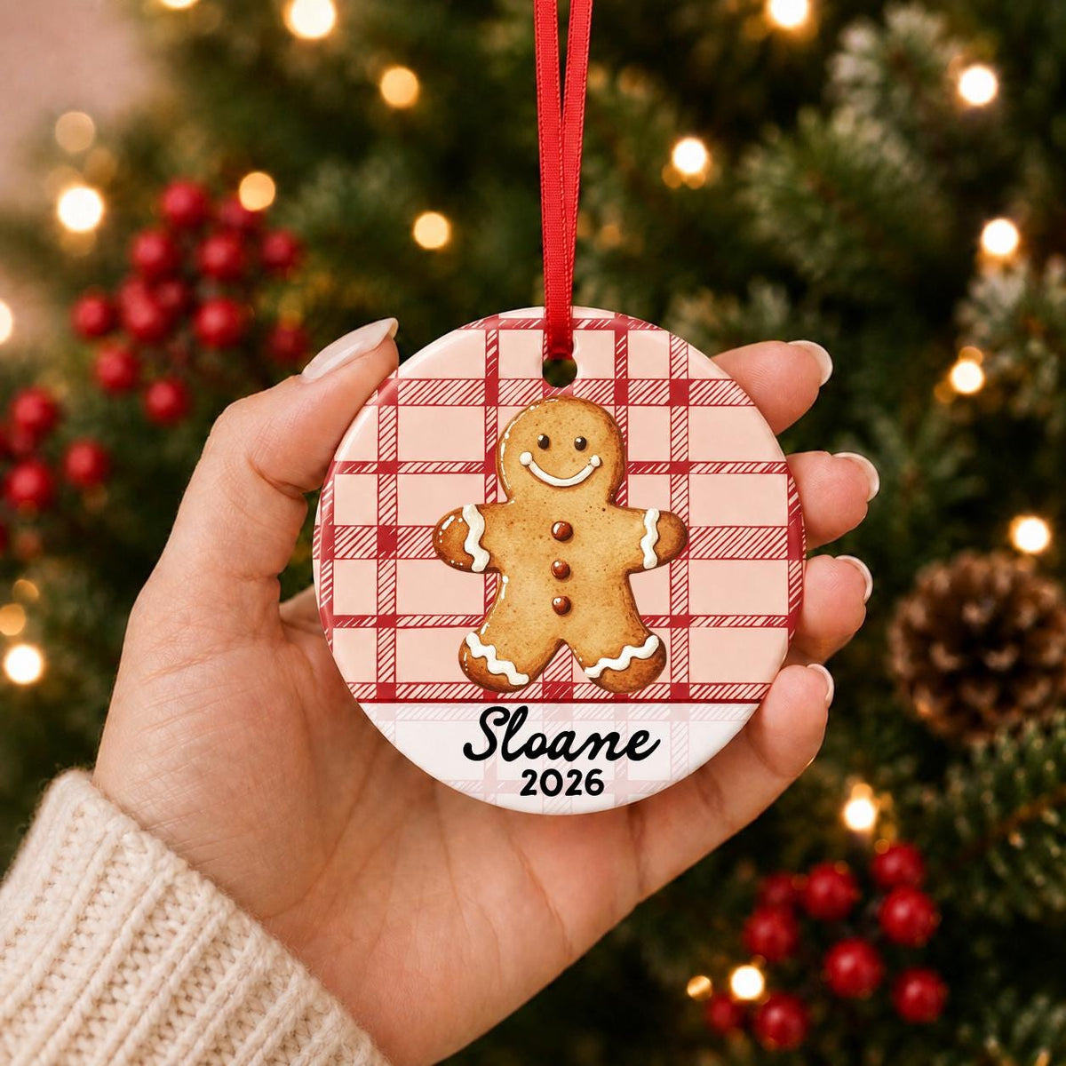 Hand holding the personalized gingerbread man ceramic ornament with name and year on red plaid in front of a Christmas tree