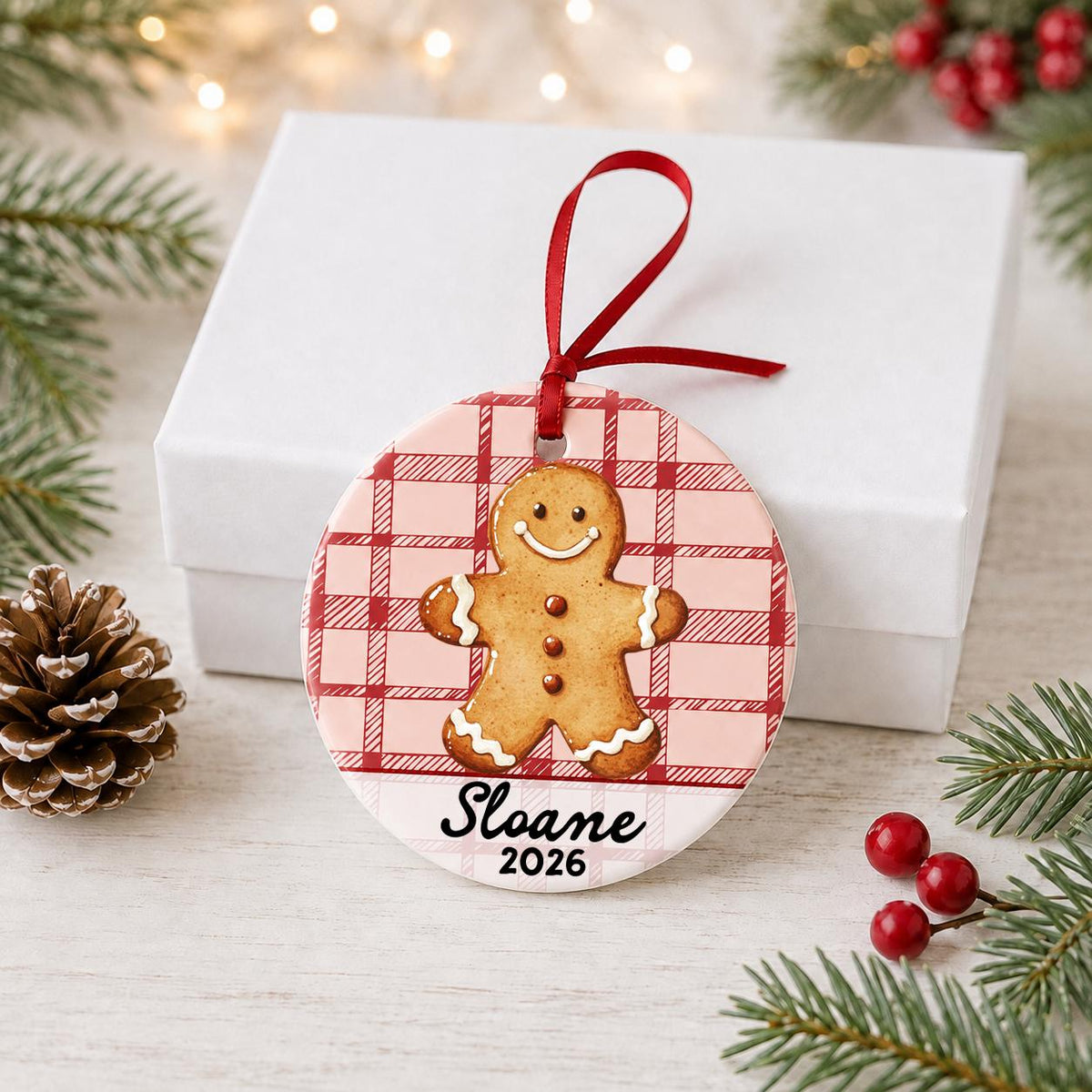 Personalized gingerbread man ceramic ornament with name and year on red plaid in front of a white gift box with pinecones and red berries
