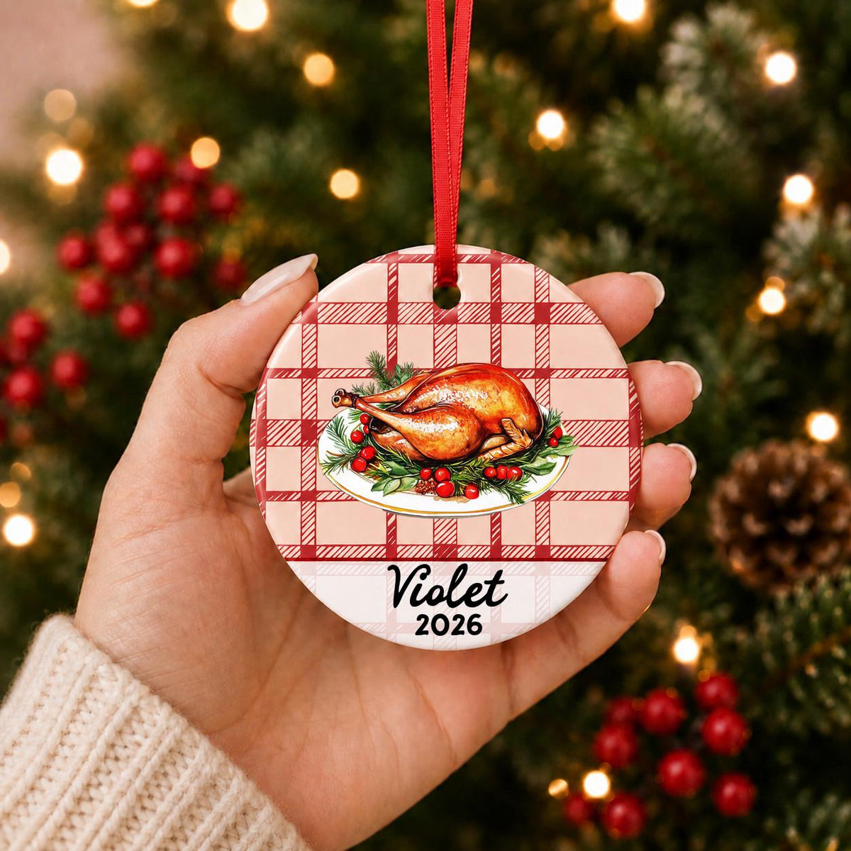 Hand holding the personalized roast turkey ceramic ornament with name and year on red plaid in front of a Christmas tree