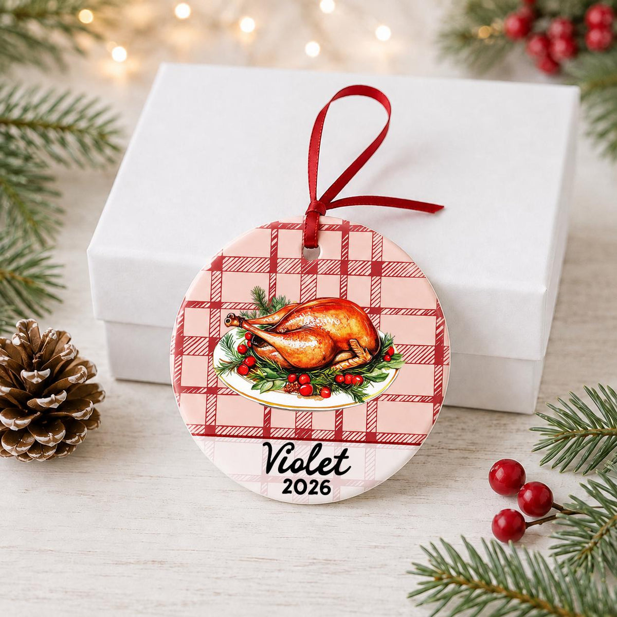 Personalized roast turkey ceramic ornament with name and year on red plaid in front of a white gift box with pinecones and red berries