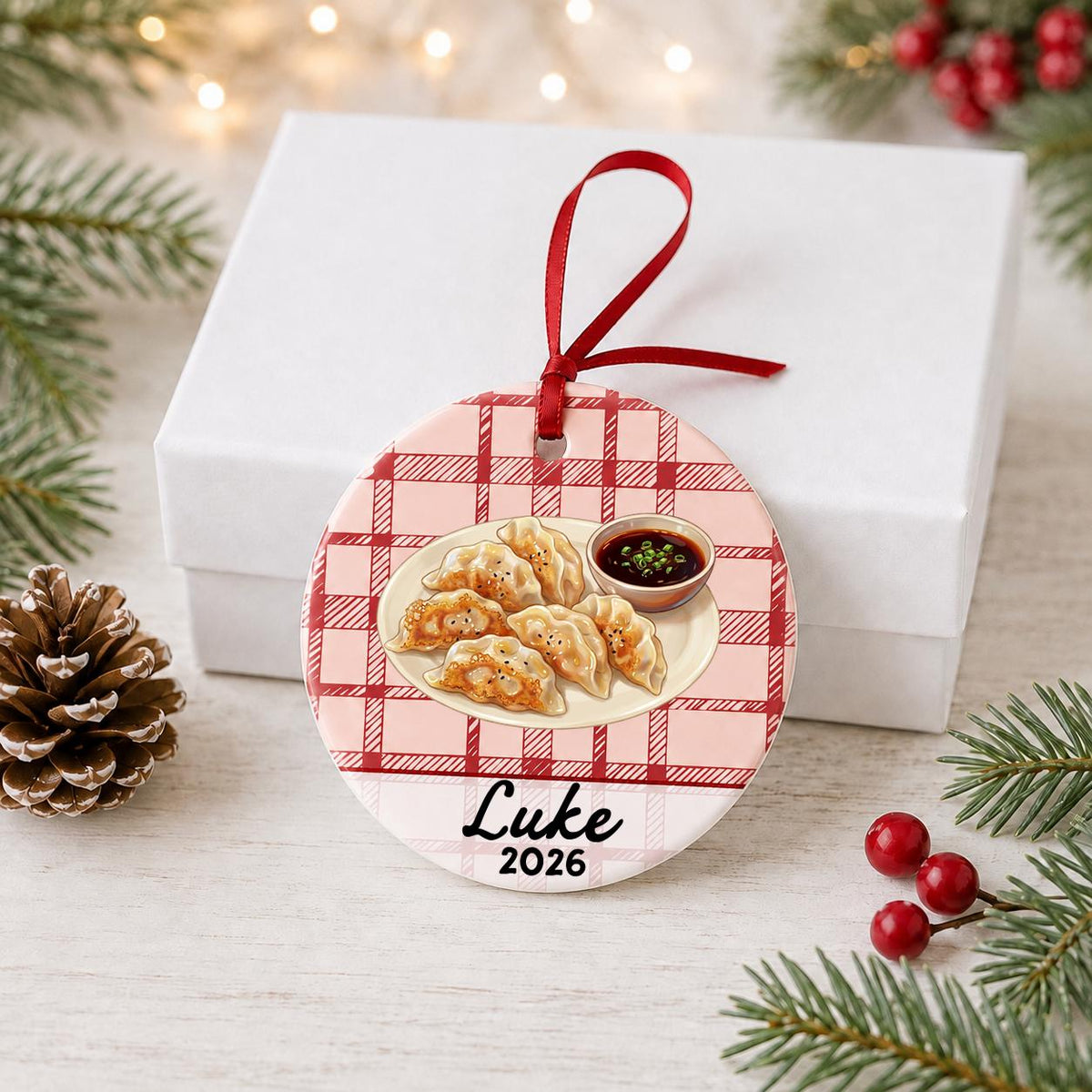 Personalized potsticker ceramic ornament with name and year on red plaid in front of a white gift box with pinecones and red berries