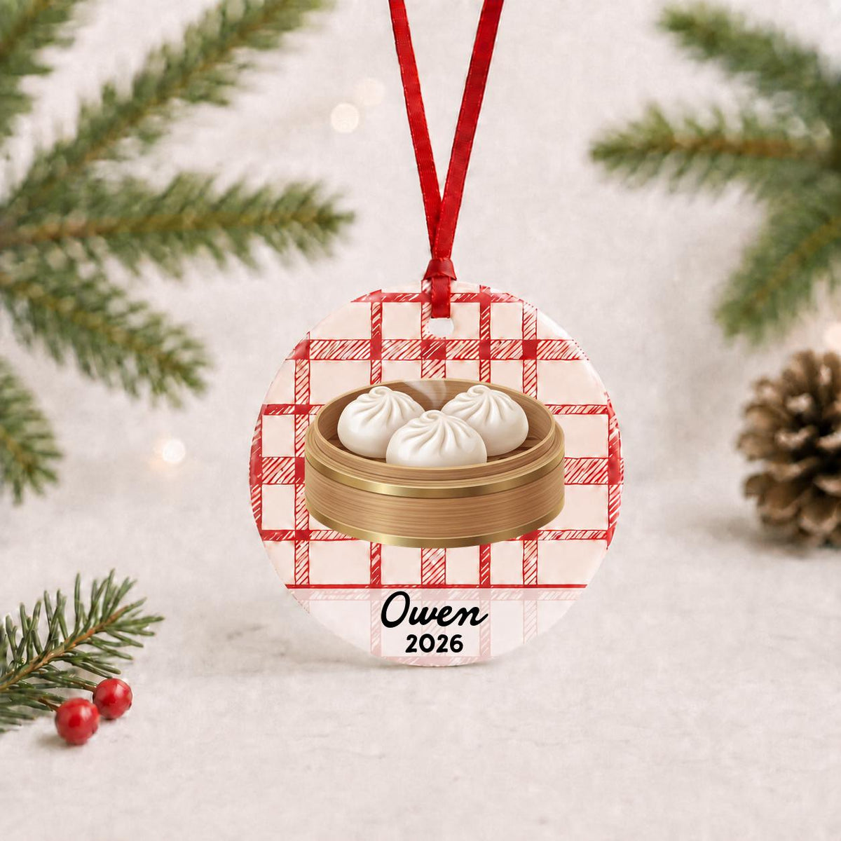 Personalized soup dumpling ceramic ornament with name and year on red plaid with red ribbon on a white surface with pine branches and pinecones
