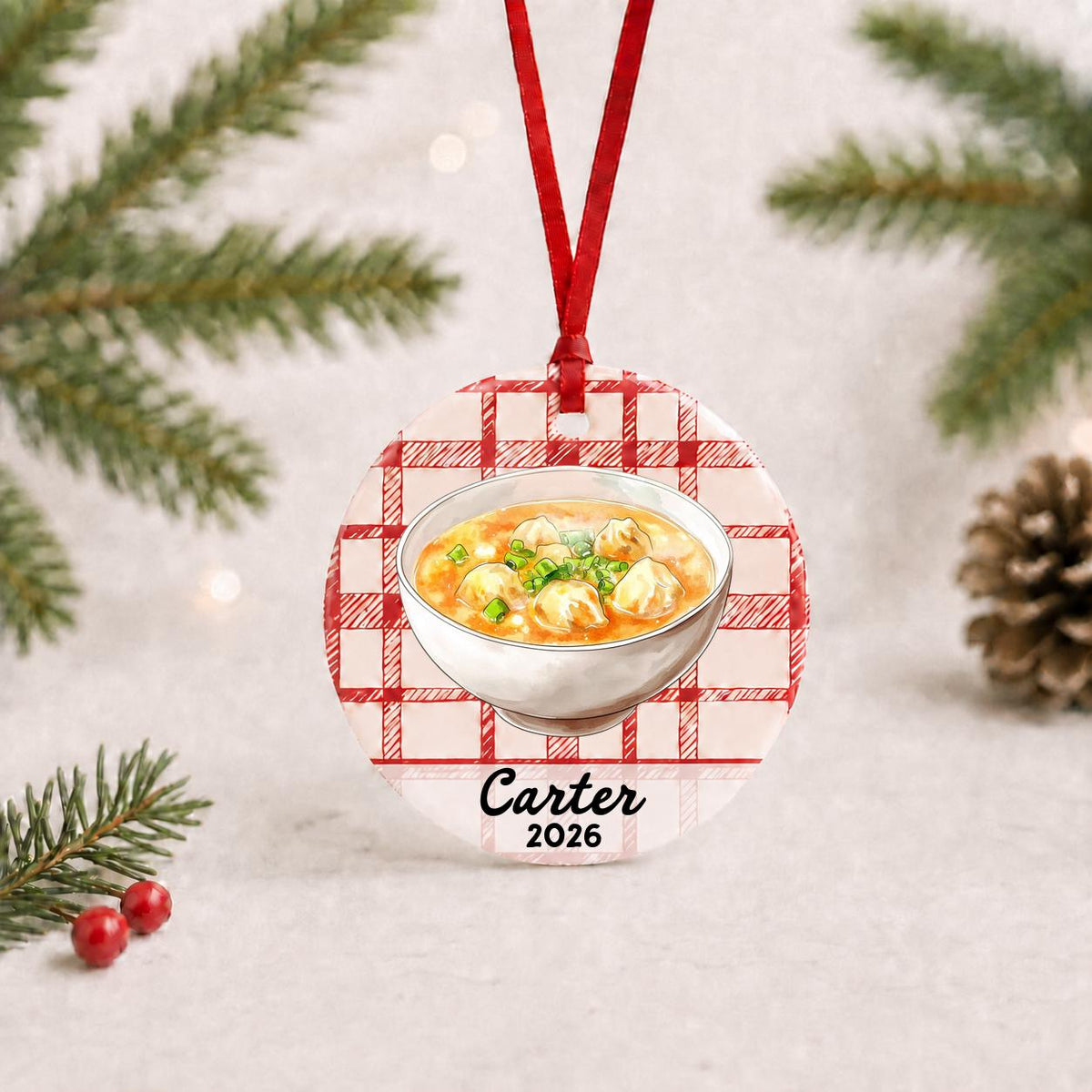 Personalized wonton soup ceramic ornament with name and year on red plaid with red ribbon on a white surface with pine branches and pinecones