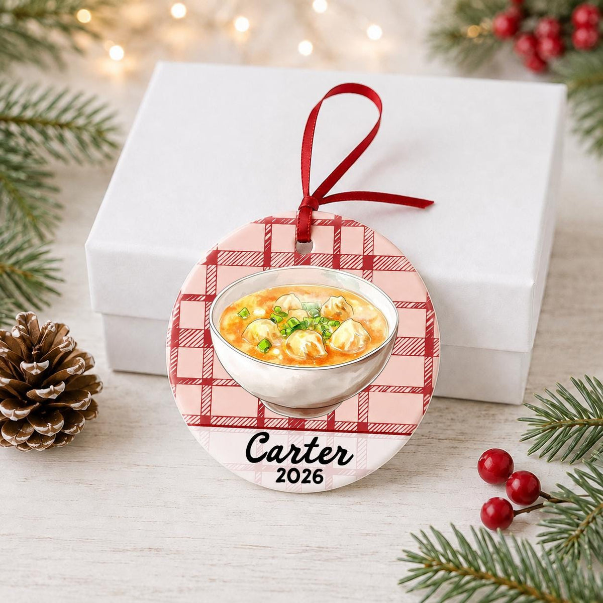 Personalized wonton soup ceramic ornament with name and year on red plaid in front of a white gift box with pinecones and red berries