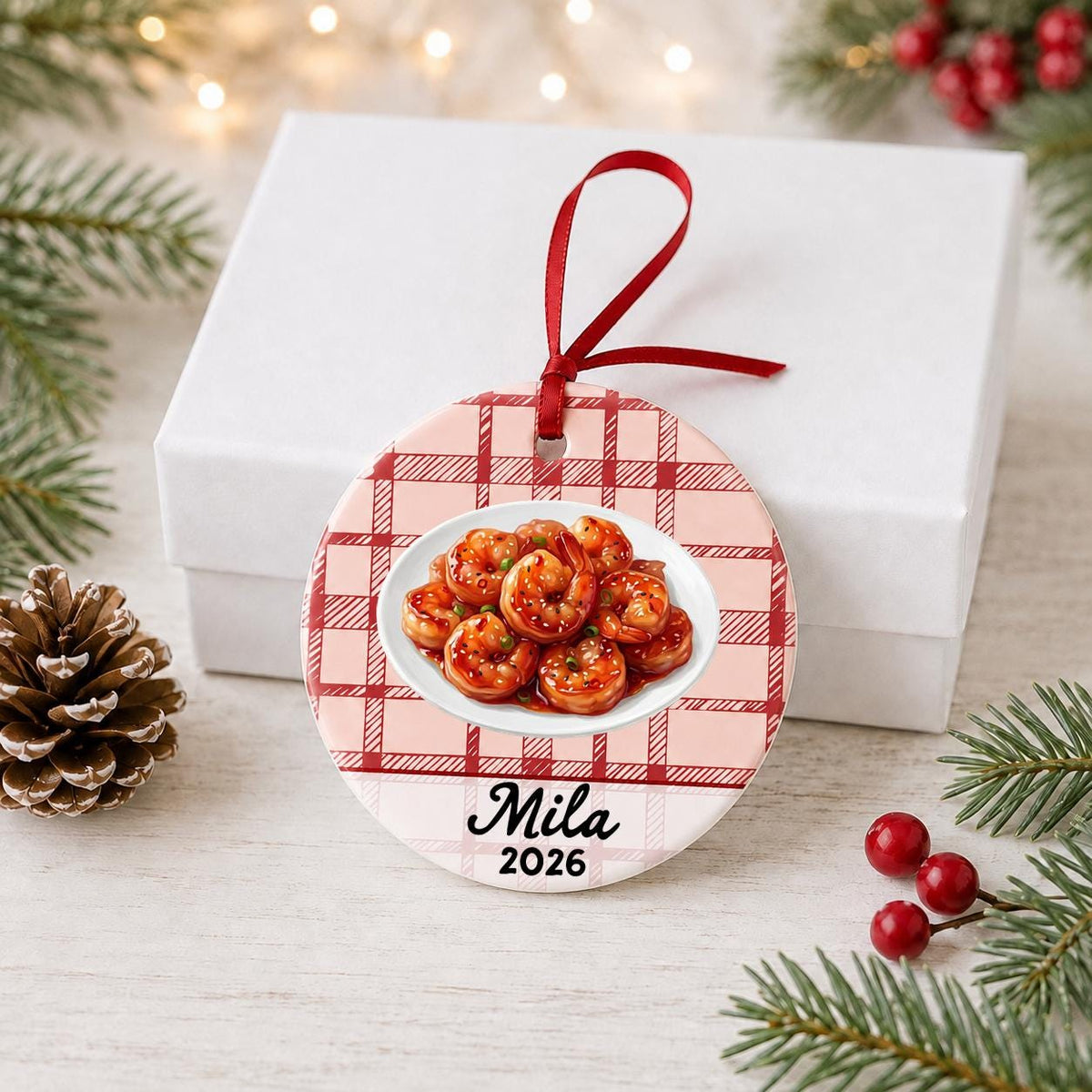 Personalized sesame shrimp ceramic ornament with name and year on red plaid in front of a white gift box with pinecones and red berries