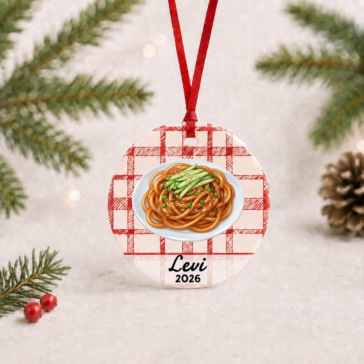 Personalized sesame noodles ceramic ornament with name and year on red plaid with red ribbon on a white surface with pine branches and pinecones