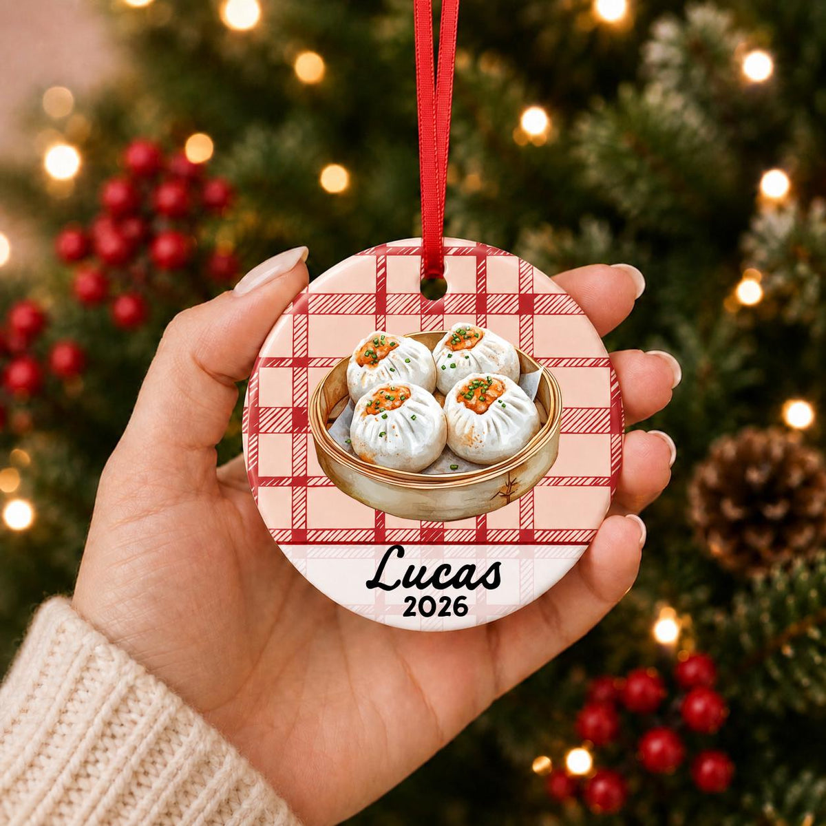 Hand holding the personalized shumai ceramic ornament with name and year on red plaid in front of a Christmas tree