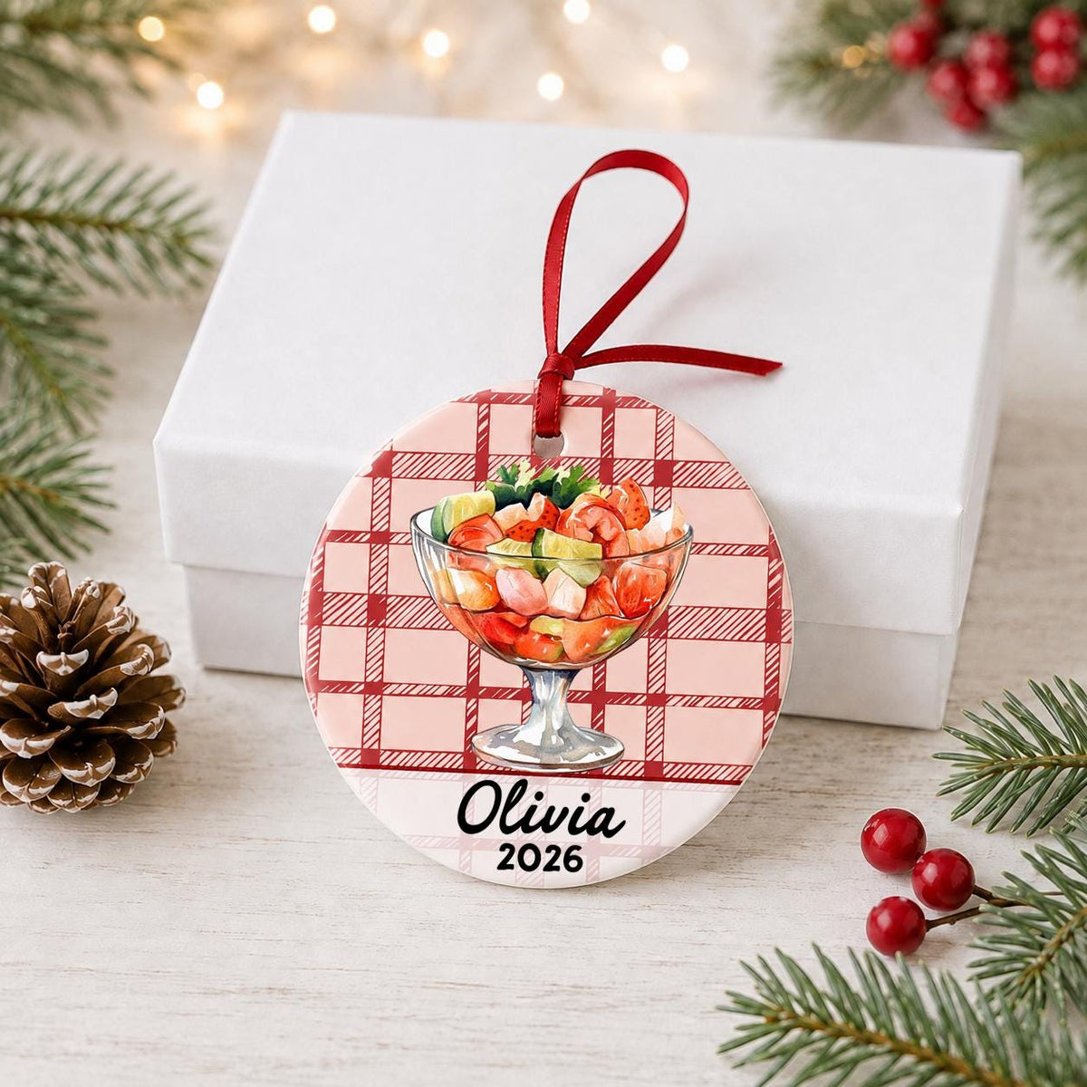 Personalized shrimp cocktail ceramic ornament with name and year on red plaid in front of a white gift box with pinecones and red berries