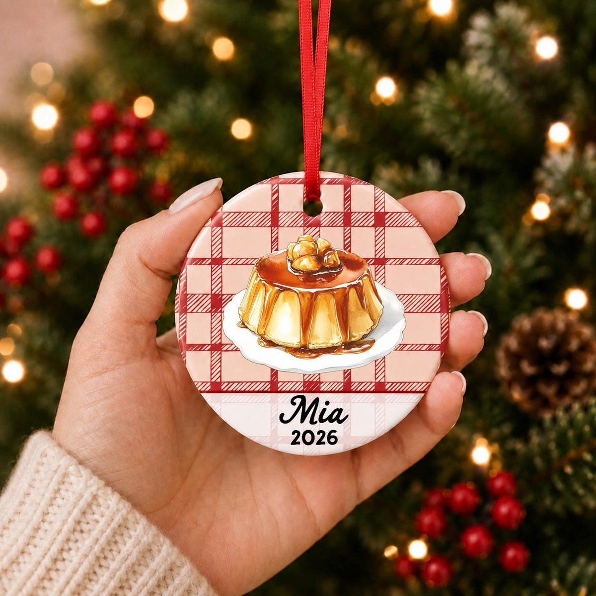 Hand holding the personalized flan ceramic ornament with name and year on red plaid in front of a Christmas tree