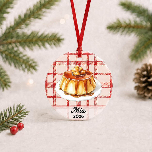 Personalized flan ceramic ornament with name and year on red plaid with red ribbon on a white surface with pine branches and pinecones