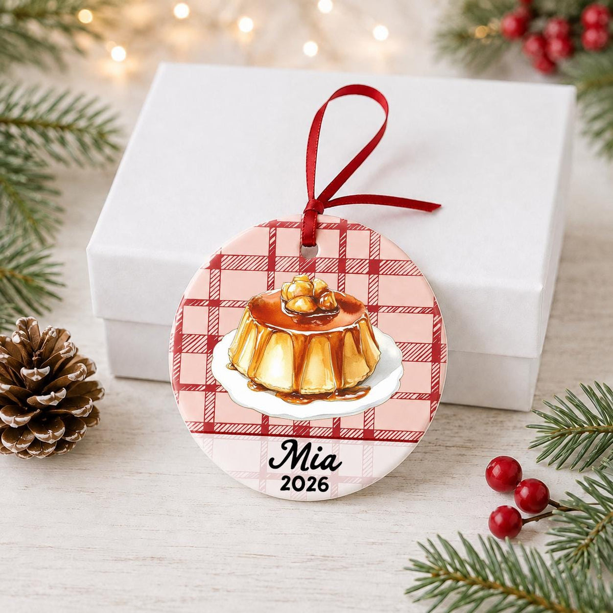 Personalized flan ceramic ornament with name and year on red plaid in front of a white gift box with pinecones and red berries