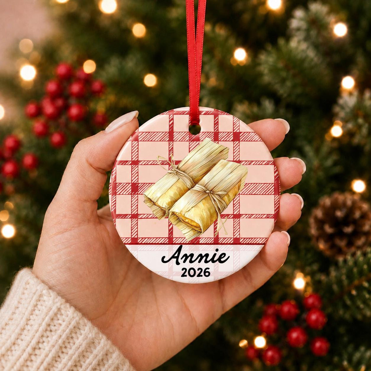 Hand holding the personalized tamale ceramic ornament with name and year on red plaid in front of a Christmas tree