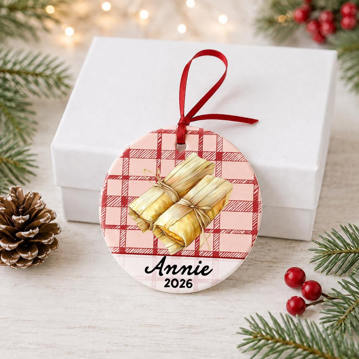 Personalized tamale ceramic ornament with name and year on red plaid in front of a white gift box with pinecones and red berries