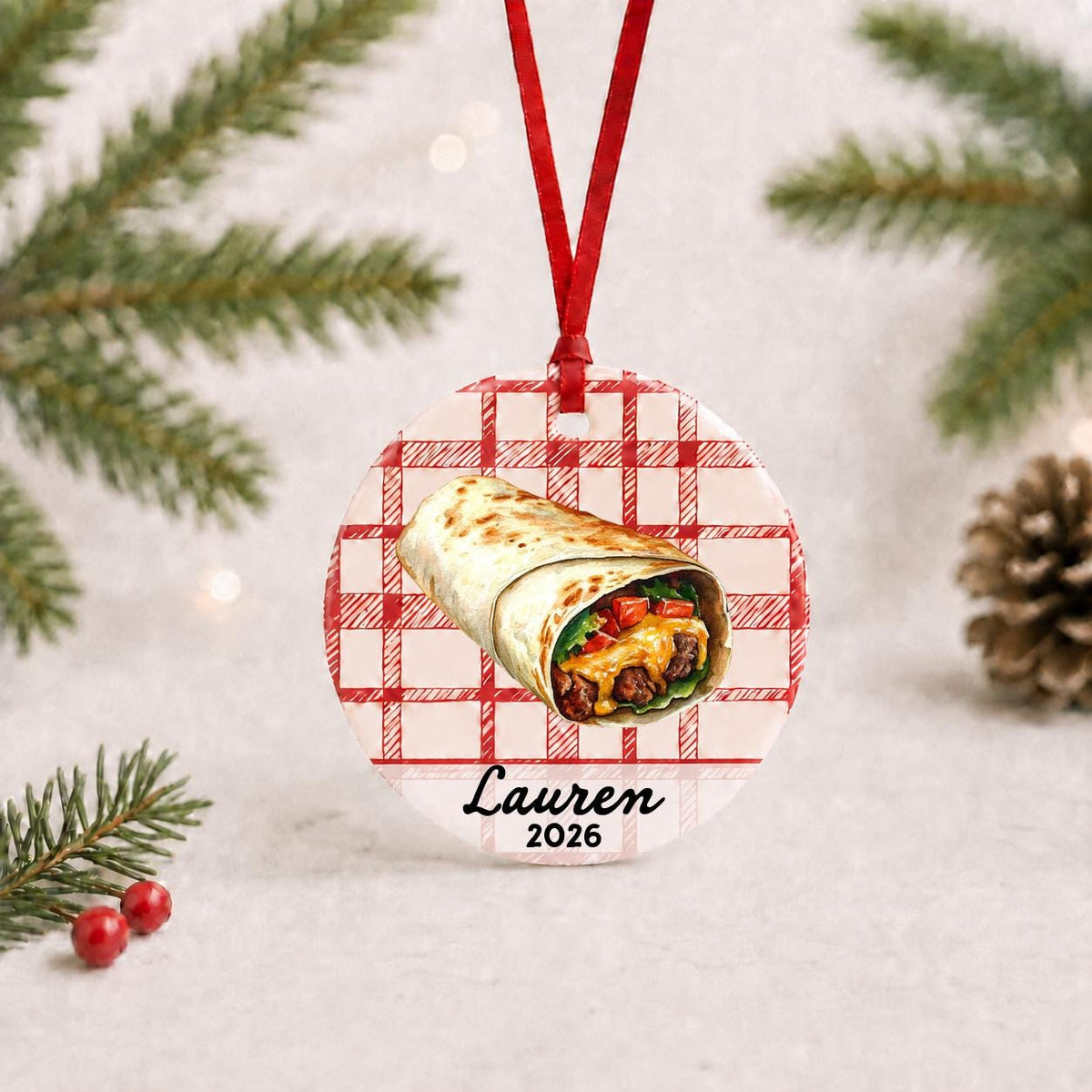 Personalized burrito ceramic ornament with name and year on red plaid with red ribbon on a white surface with pine branches and pinecones