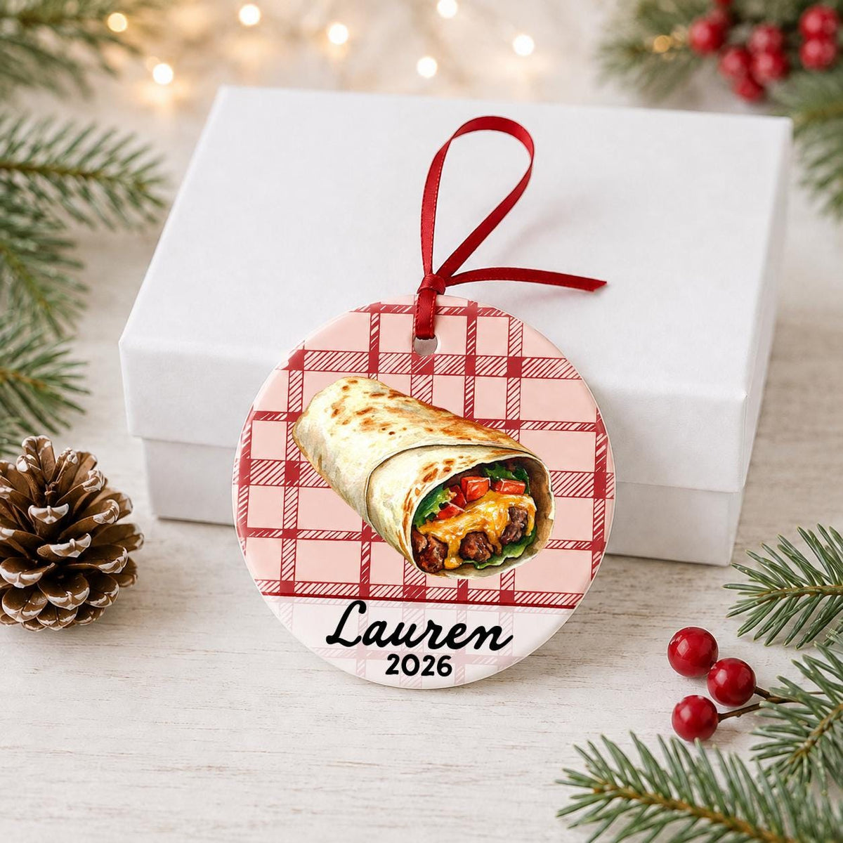 Personalized burrito ceramic ornament with name and year on red plaid in front of a white gift box with pinecones and red berries