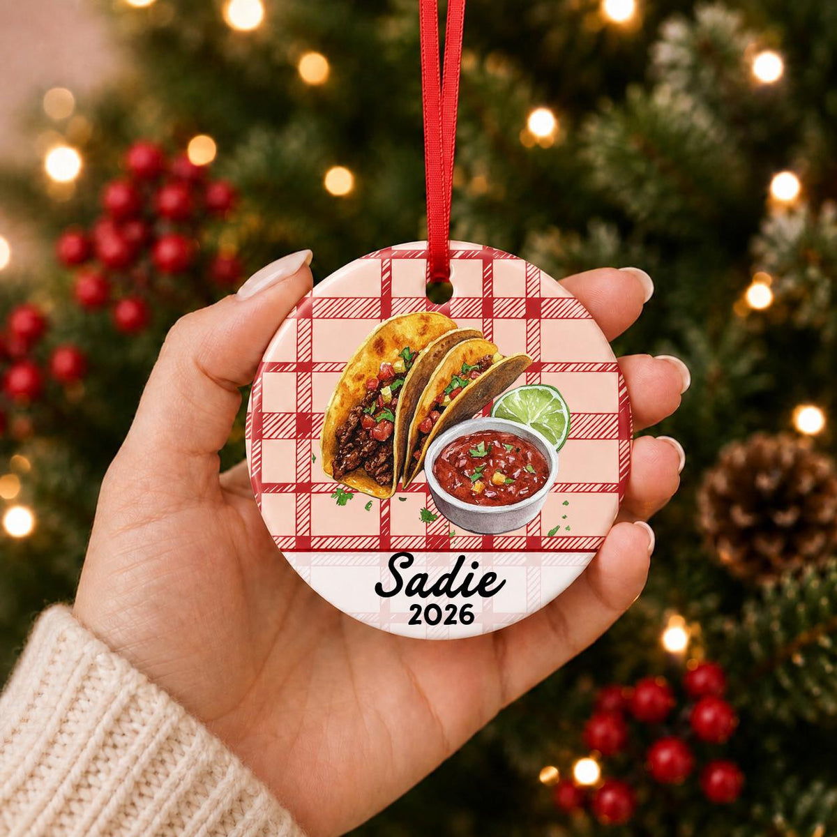 Hand holding the personalized taco ceramic ornament with name and year on red plaid in front of a Christmas tree