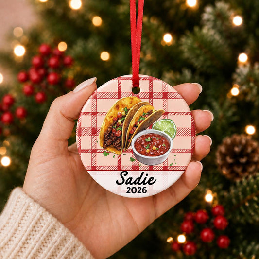 Hand holding the personalized taco ceramic ornament with name and year on red plaid in front of a Christmas tree