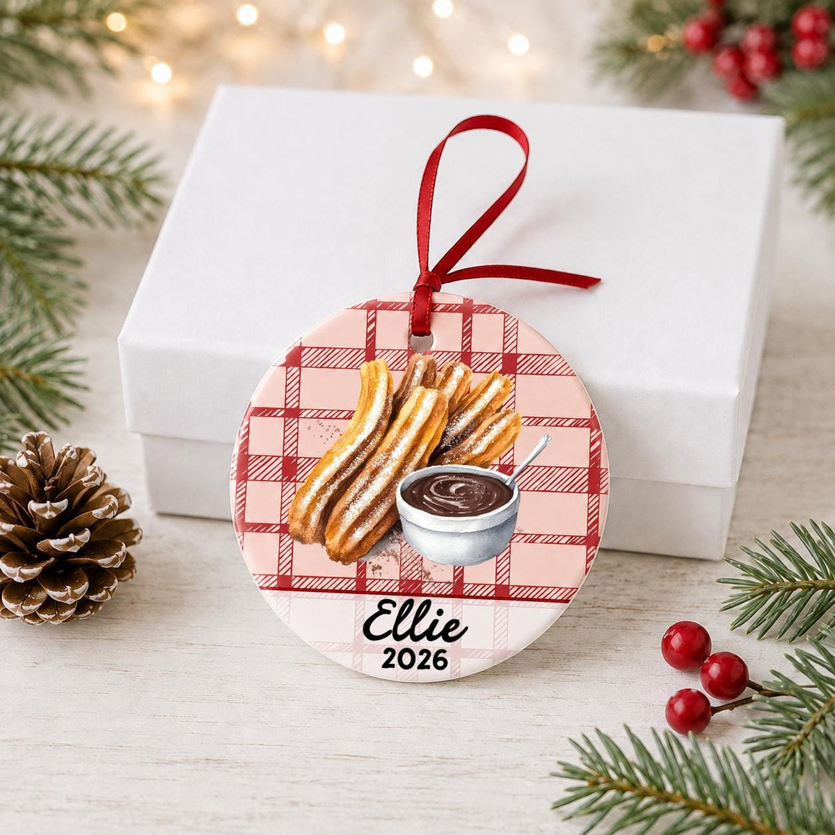 Personalized churro ceramic ornament with name and year on red plaid in front of a white gift box with pinecones and red berries