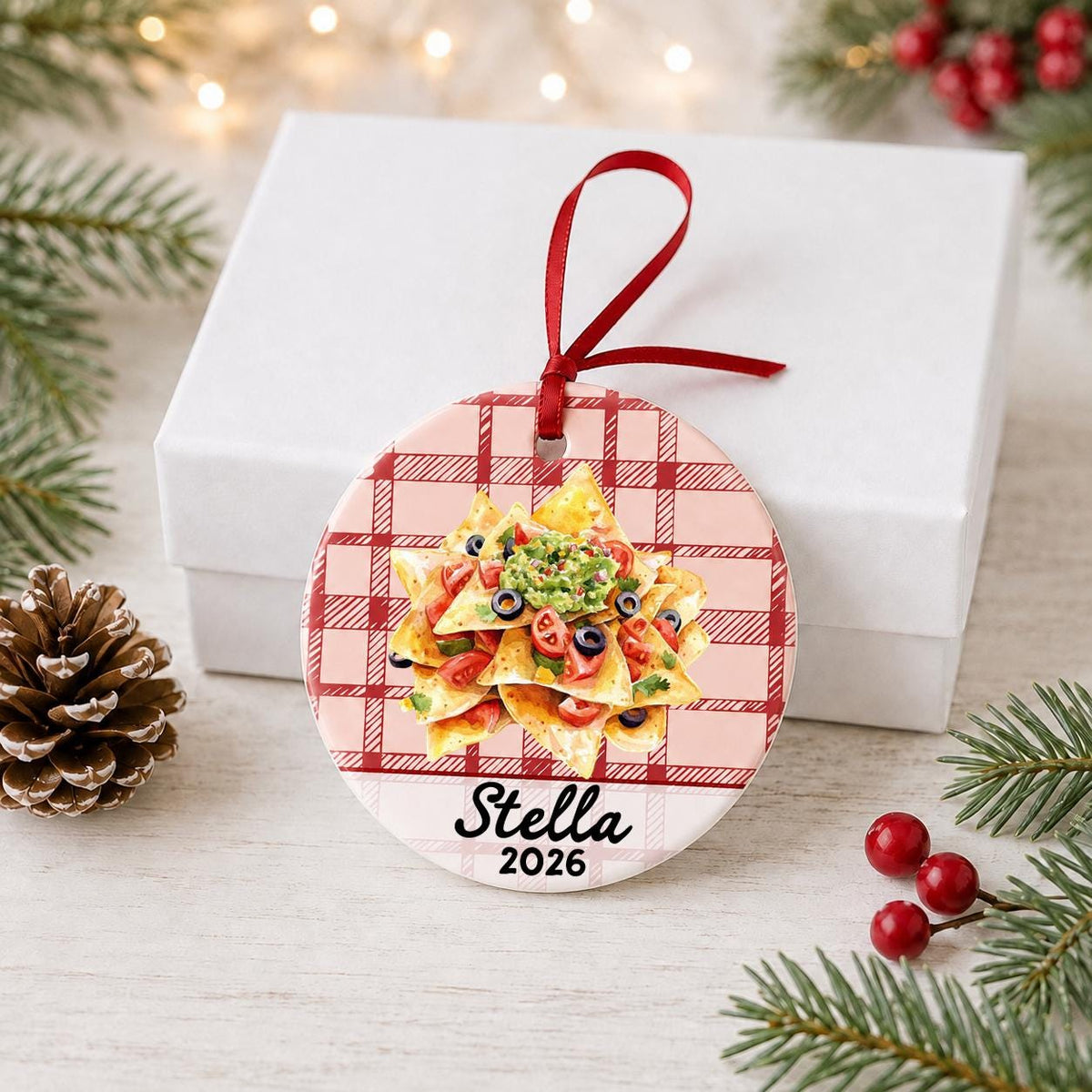 Personalized nachos ceramic ornament with name and year on red plaid in front of a white gift box with pinecones and red berries