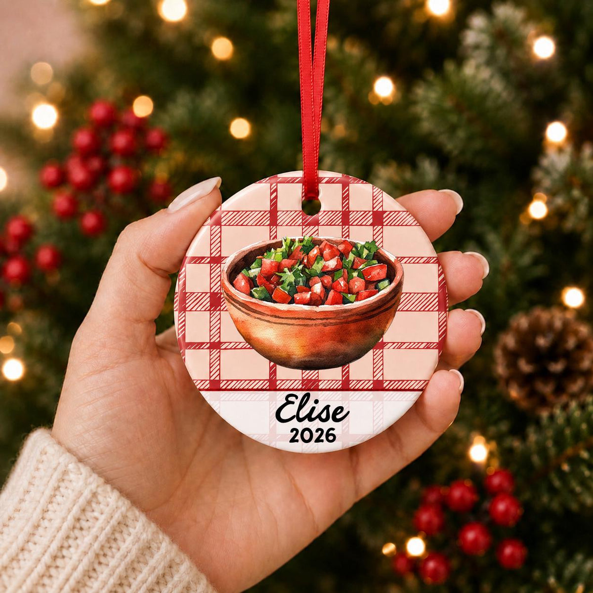 Hand holding the personalized pico de gallo ceramic ornament with name and year on red plaid in front of a Christmas tree
