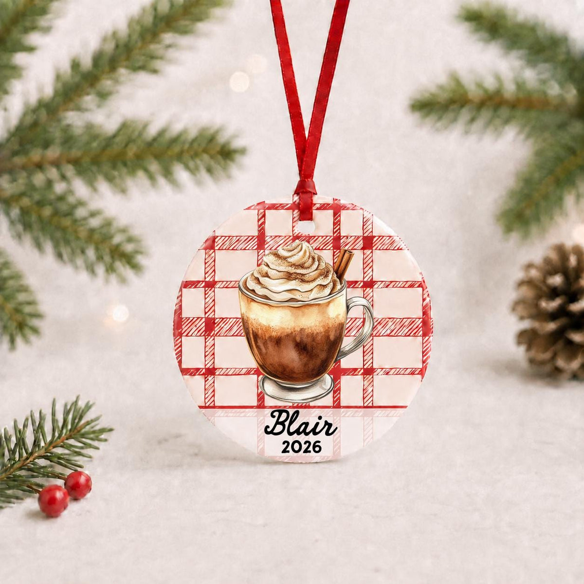 Personalized Mexican hot chocolate ceramic ornament with name and year on red plaid with red ribbon on a white surface with pine branches and pinecones