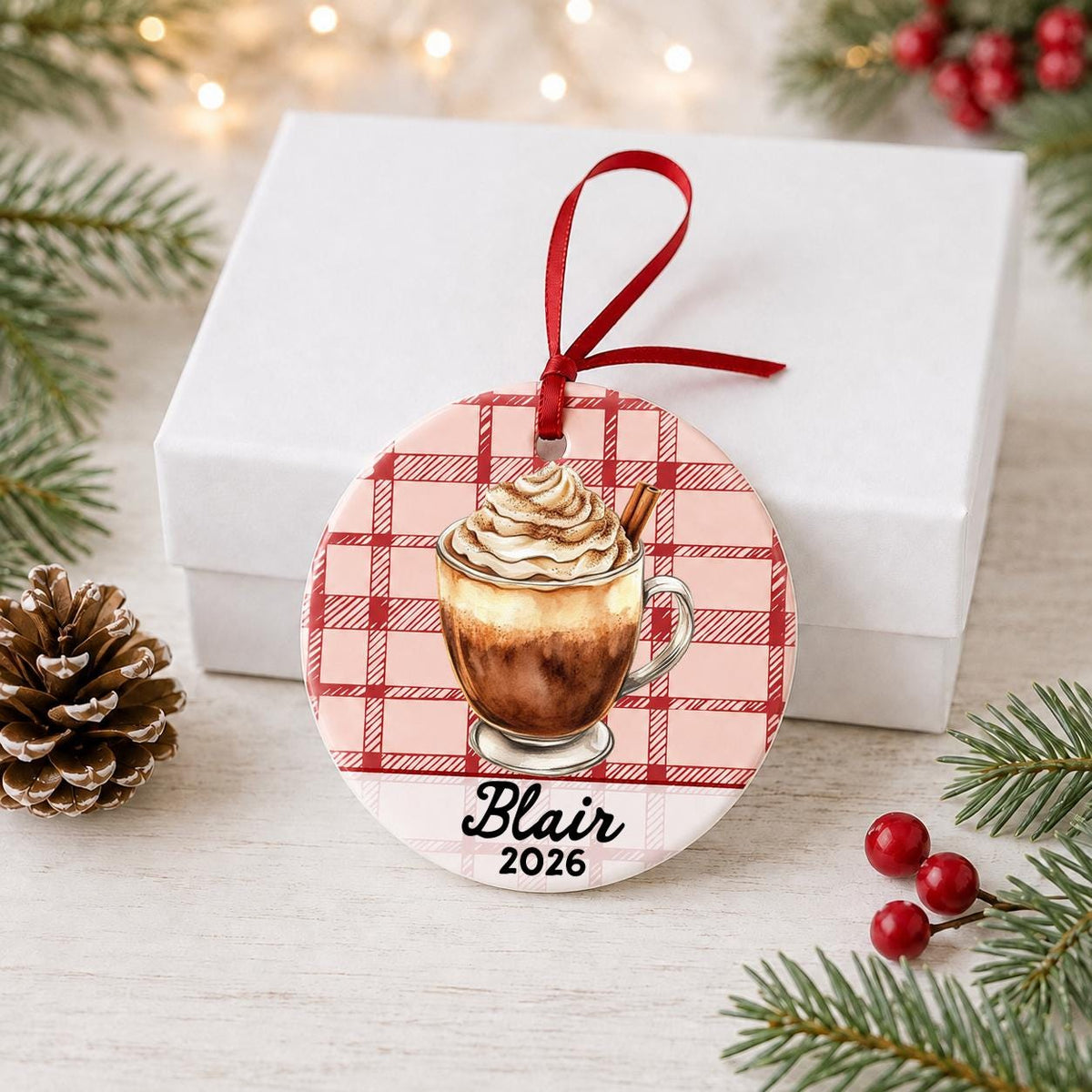Personalized Mexican hot chocolate ceramic ornament with name and year on red plaid in front of a white gift box with pinecones and red berries