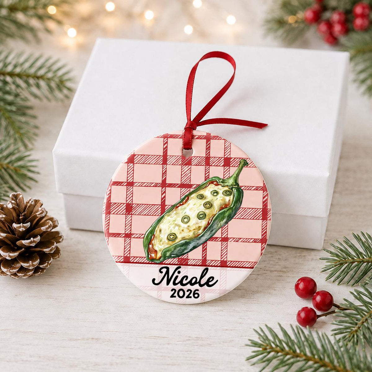 Personalized chile relleno ceramic ornament with name and year on red plaid in front of a white gift box with pinecones and red berries
