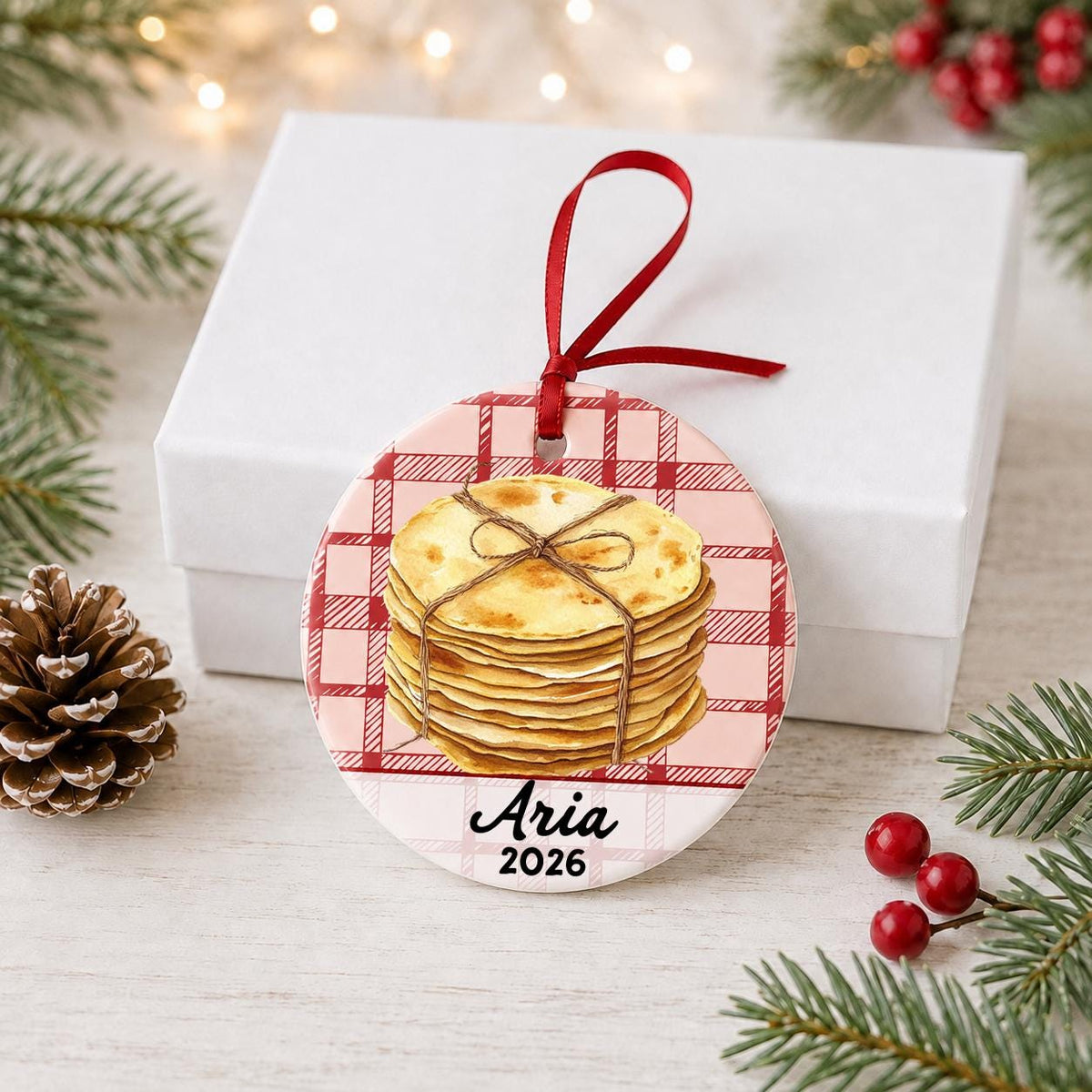 Personalized tortilla ceramic ornament with name and year on red plaid in front of a white gift box with pinecones and red berries