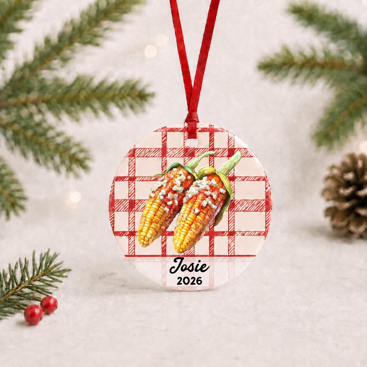 Personalized Mexican street corn ceramic ornament with name and year on red plaid with red ribbon on a white surface with pine branches and pinecones
