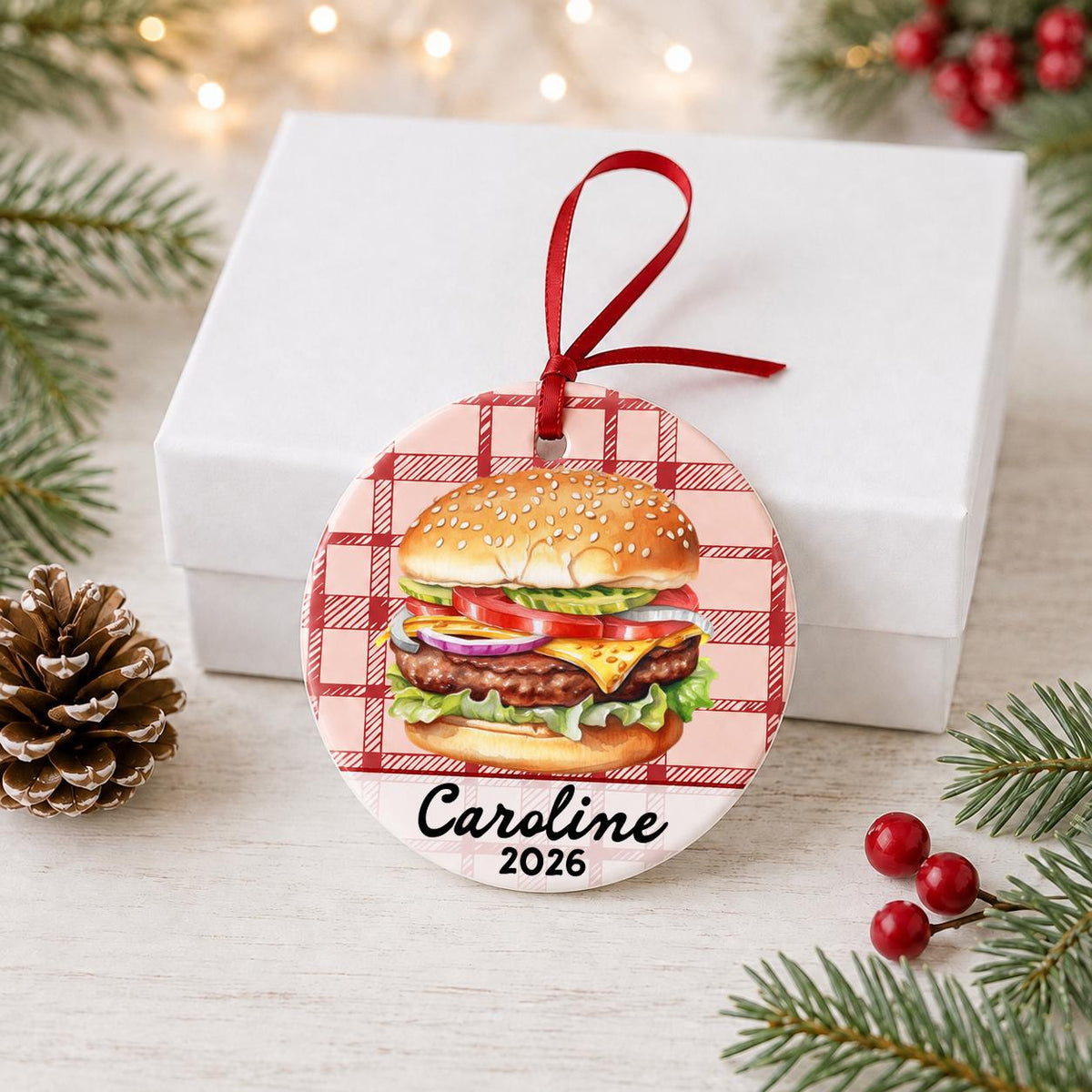 Personalized cheeseburger ceramic ornament with name and year on red plaid in front of a white gift box with pinecones and red berries