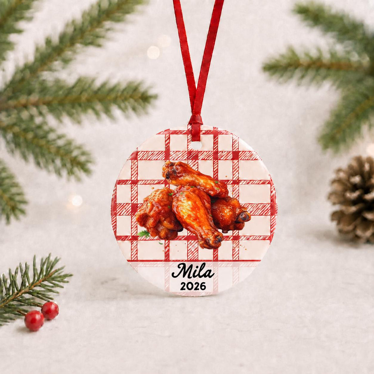 Personalized buffalo wings ceramic ornament with name and year on red plaid with red ribbon on a white surface with pine branches and pinecones