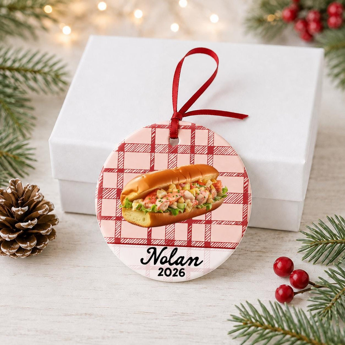 Personalized lobster roll ceramic ornament with name and year on red plaid in front of a white gift box with pinecones and red berries
