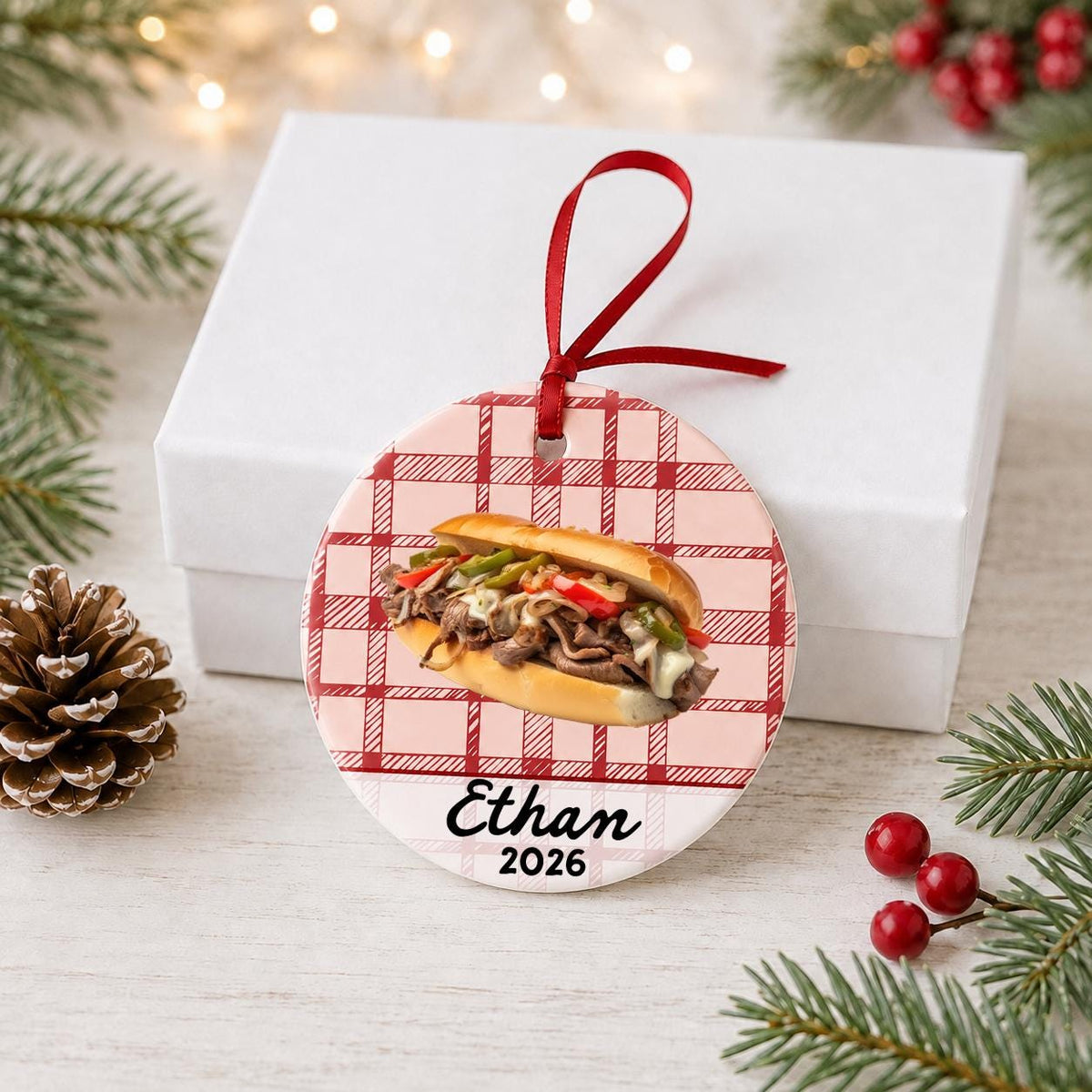 Personalized Philly cheesesteak ceramic ornament with name and year on red plaid in front of a white gift box with pinecones and red berries
