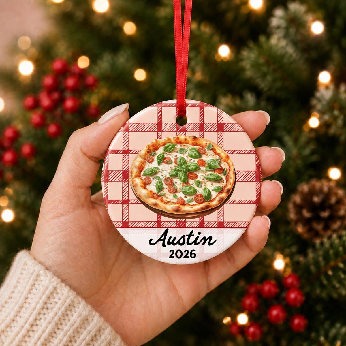 Hand holding the personalized Margherita pizza ceramic ornament with name and year on red plaid in front of a Christmas tree