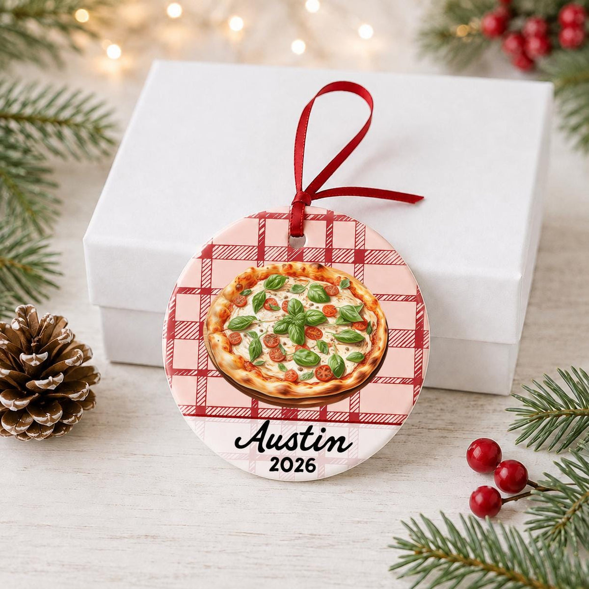 Personalized Margherita pizza ceramic ornament with name and year on red plaid in front of a white gift box with pinecones and red berries