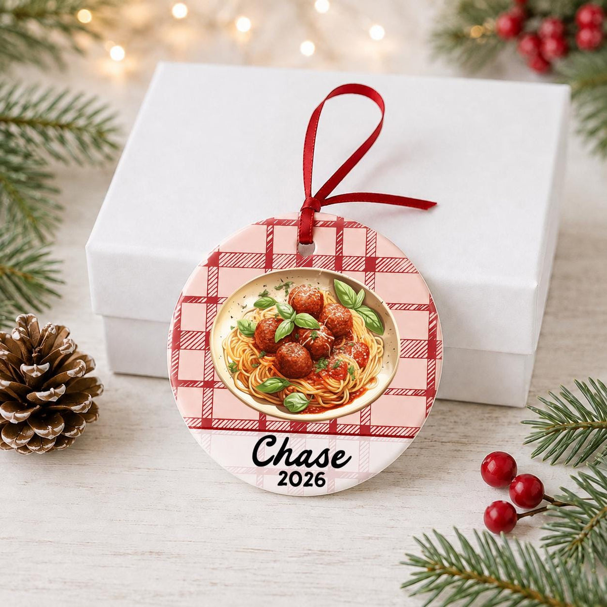 Personalized spaghetti and meatballs ceramic ornament with name and year on red plaid in front of a white gift box with pinecones and red berries