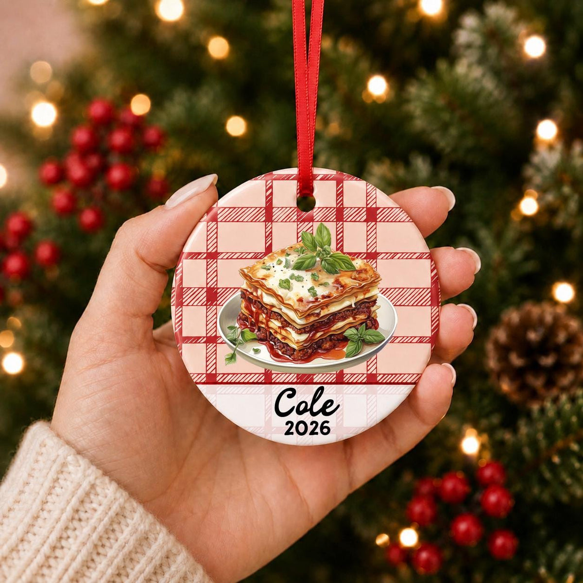 Hand holding the personalized lasagna ceramic ornament with name and year on red plaid in front of a Christmas tree