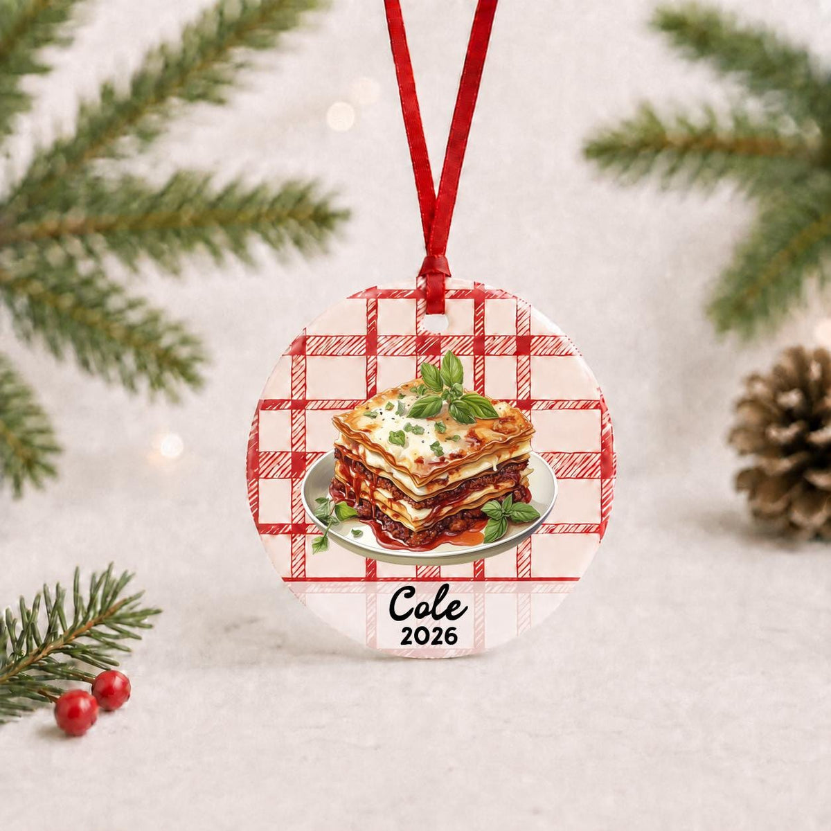 Personalized lasagna ceramic ornament with name and year on red plaid with red ribbon on a white surface with pine branches and pinecones