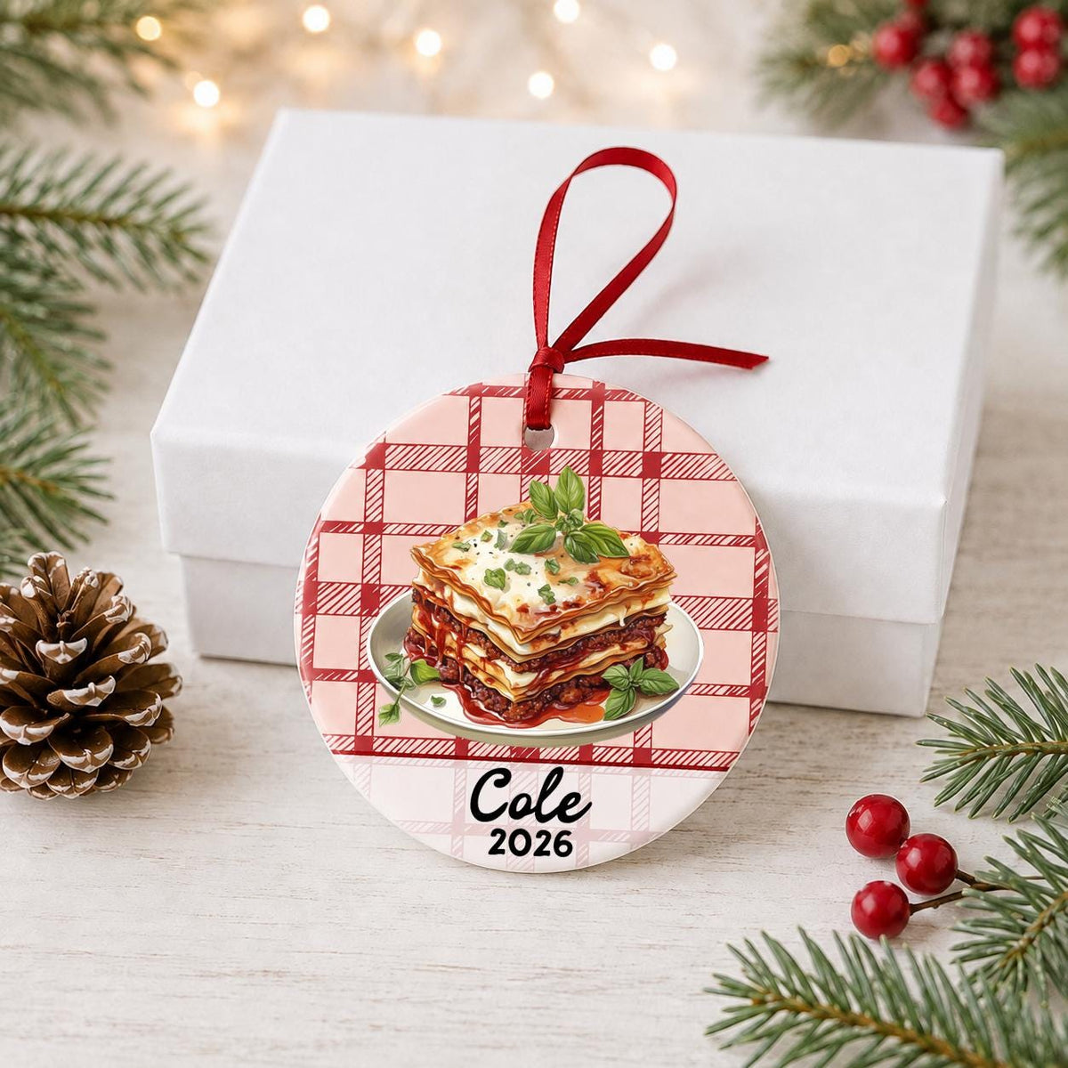 Personalized lasagna ceramic ornament with name and year on red plaid in front of a white gift box with pinecones and red berries