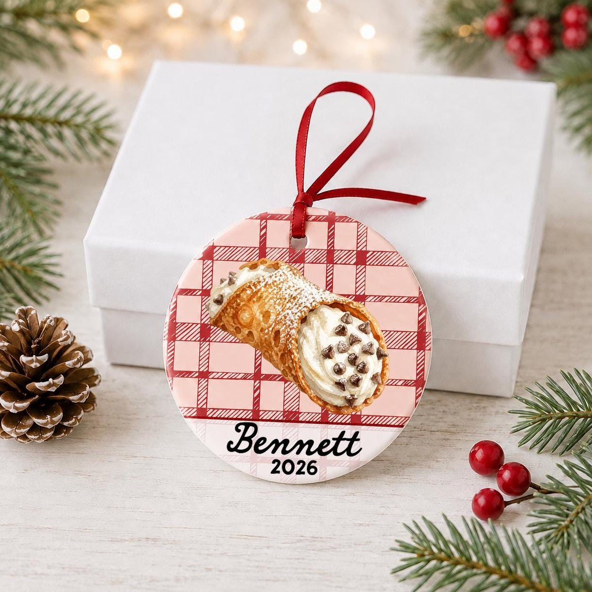 Personalized cannoli ceramic ornament with name and year on red plaid in front of a white gift box with pinecones and red berries